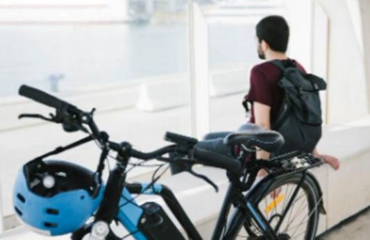 Electric bike with blue helmet in foreground, man with backpack seated by waterfront