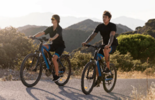 two people riding blue electric bikes on a road in a mountain landscape, side view